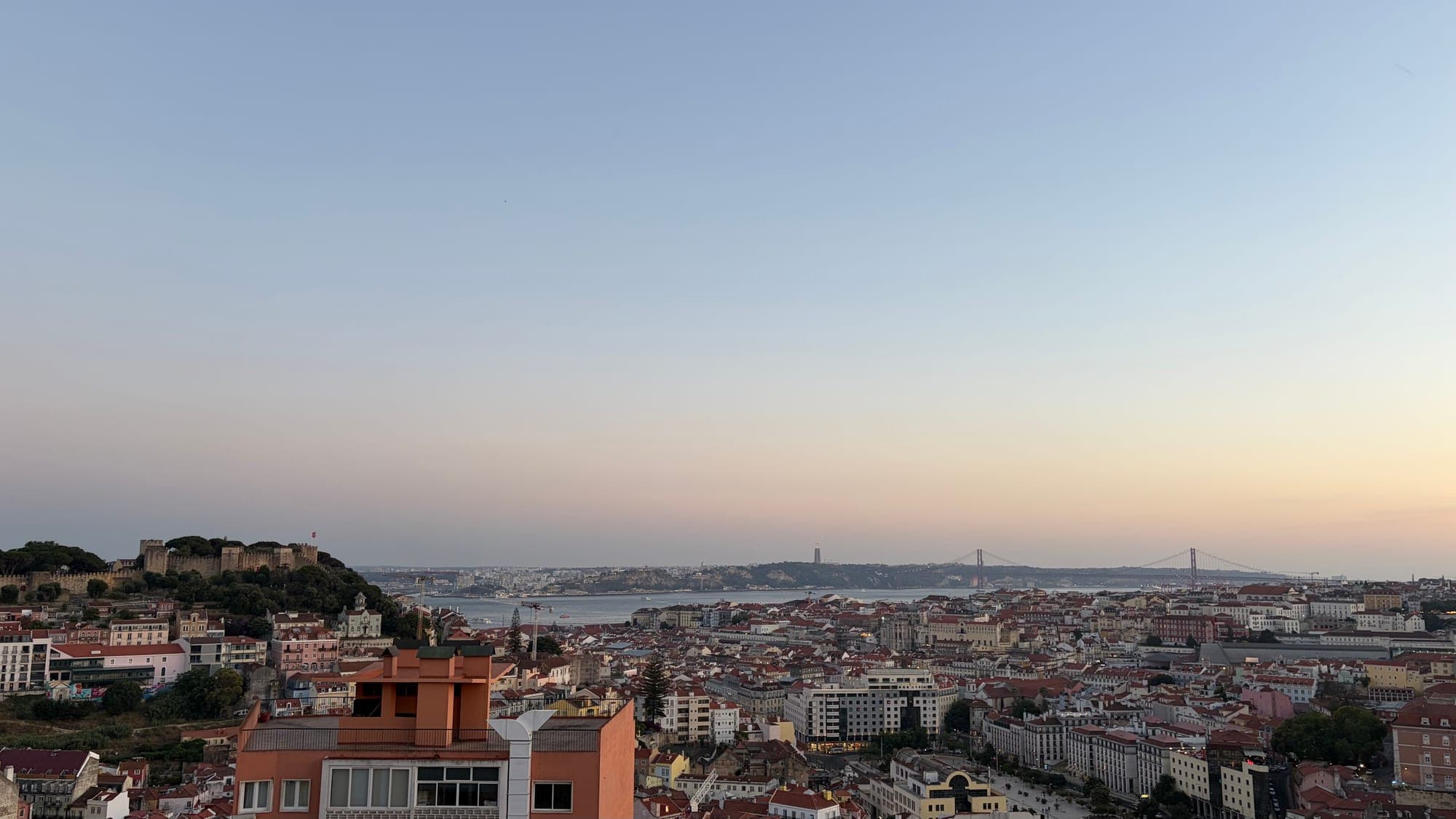 Lisboa vista de um miradouro ao entardecer, com o Castelo de São Jorge e a Ponte 25 de Abril ao fundo